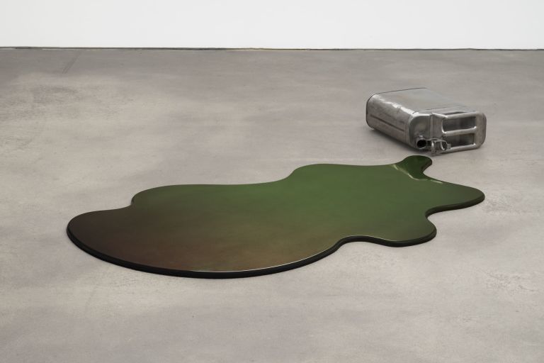 Floor sculpture: overturned silver metal jerrycan spilling an iridescent dark green-brown puddle on concrete floor.