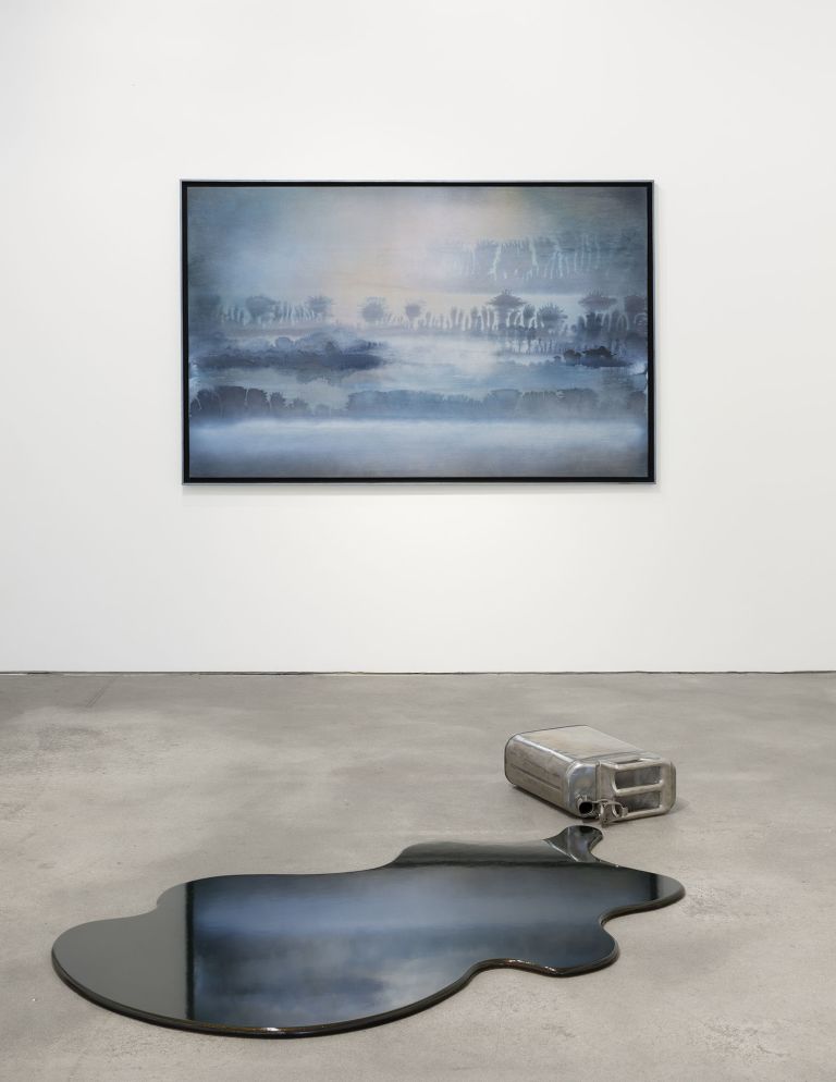 Floor sculpture: overturned silver metal jerrycan spilling an iridescent dark green-brown puddle on concrete floor.