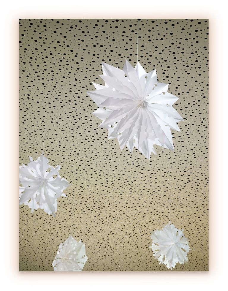 Photograph of white paper star ornaments hanging against spotted beige ceiling, varying sizes.