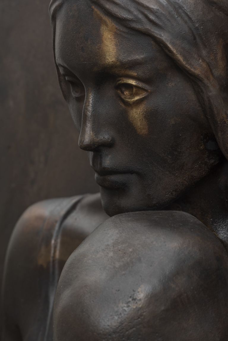 Dark bronze wall relief: seated female figure with elongated limbs, one gilded finger, on oxidized background.