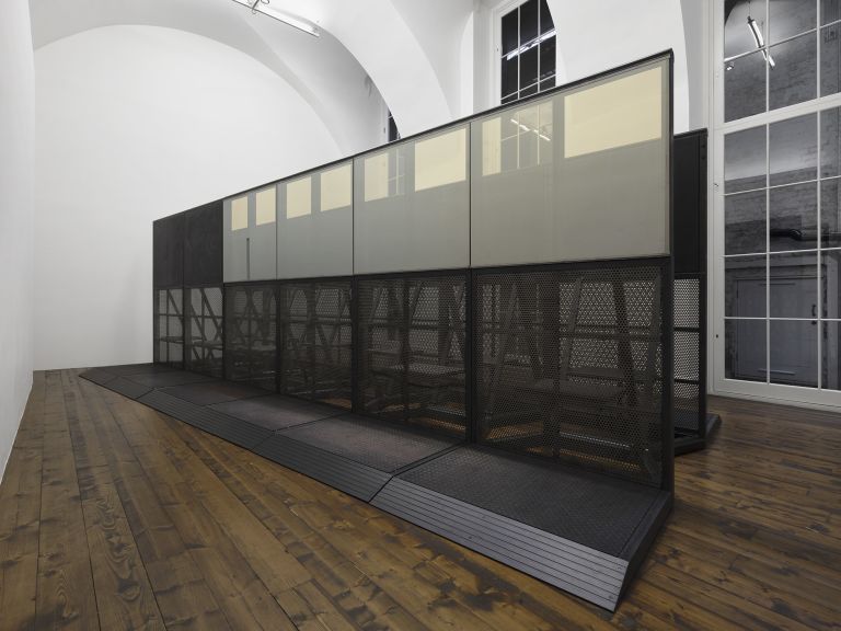 Large black metal installation: modular structure with perforated mesh lower panels and grey-tinted glass upper panels on a platform.