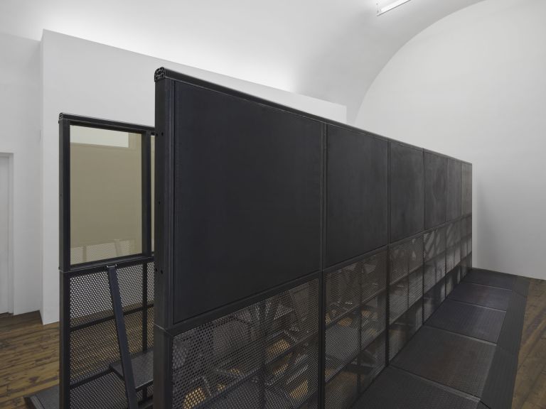 Large black metal installation: modular structure with perforated mesh lower panels and grey-tinted glass upper panels on a platform.