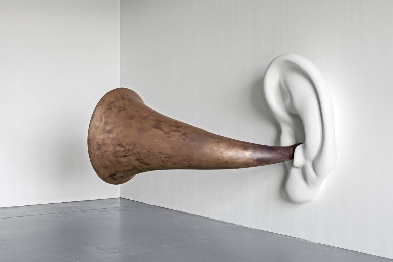 Sculptural installation of bronze horn attached to white ceramic ear mounted on wall