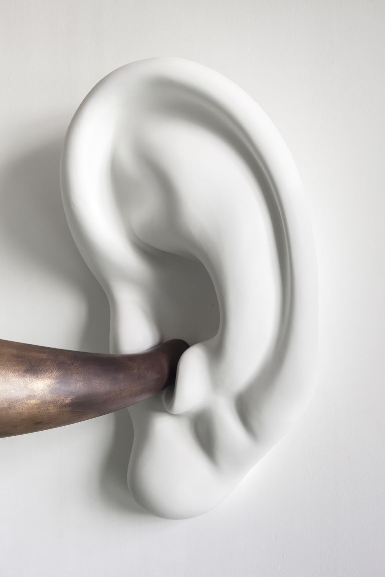 Sculptural installation of bronze horn attached to white ceramic ear mounted on wall