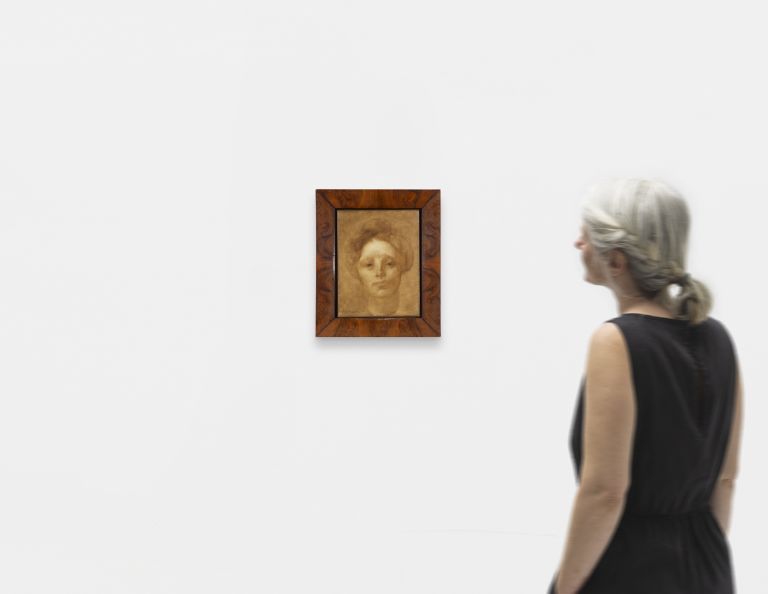 Portrait painting in ornate wooden frame showing a person with curly hair rendered in warm, sepia tones on canvas.
