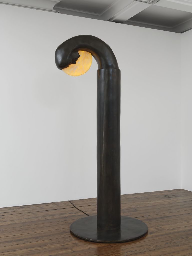 Close-up of black curved sculptural lamp with yellow interior light, standing on circular base in gallery setting.