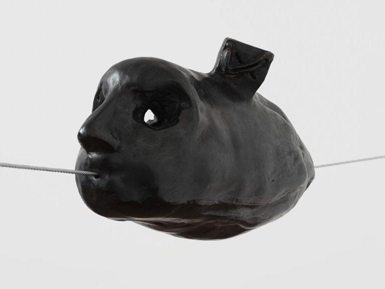 Black sculptural object resembling a head or face suspended by thin wire, with glossy ceramic-like finish.