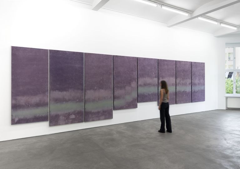 Eight purple abstract paintings arranged in a horizontal line on a white gallery wall, featuring cloudy, atmospheric textures.