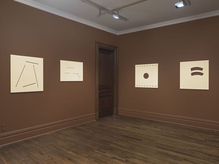 Gallery corner with brown walls and wooden floor displaying four minimalist cream-colored artworks with simple line cuts, dotted lines, and curved forms.