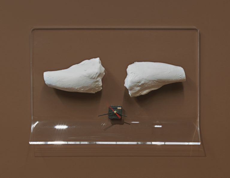 Two white sculptural fragments mounted on a clear acrylic display with a small clock mechanism between them, all set against a brown wall background.