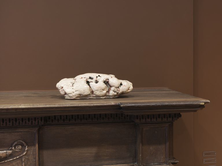 A white, lumpy sculptural object with dark spots or crevices displayed on a wooden antique mantelpiece against a brown wall.