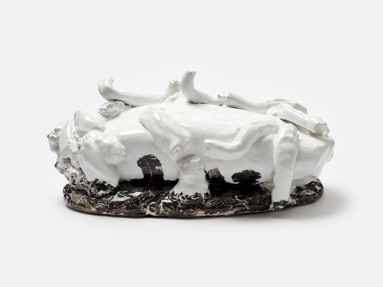 A black and white sculptural ceramic object with protruding elements resembling bones or limbs, partially resting on a black base, photographed against a white background.