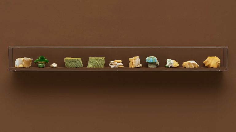 Clear acrylic shelf mounted on brown wall displaying a collection of small foam objects in green, cream, and blue tones.