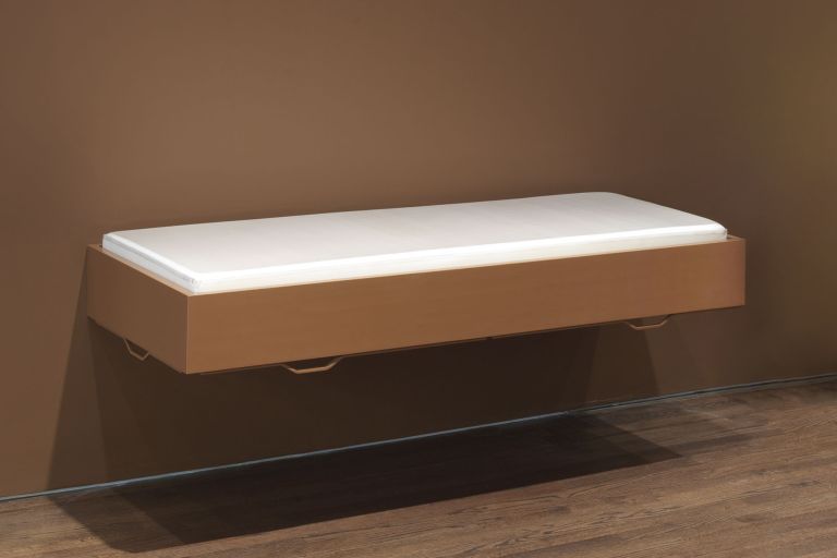 Wall-mounted floating bed frame in medium brown wood with white mattress, featuring angular metal handles at corners, displayed against plain white wall in gallery setting.