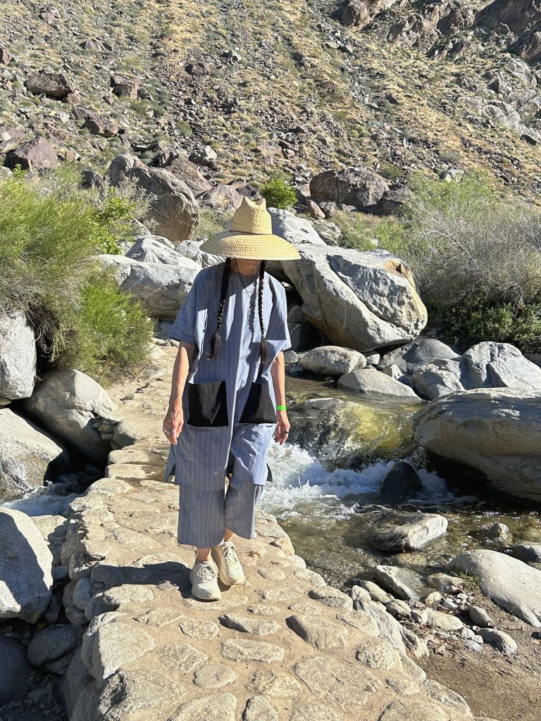 Figure in hat and casual clothing walking on rocky path through desert landscape with scattered boulders and sparse vegetation.