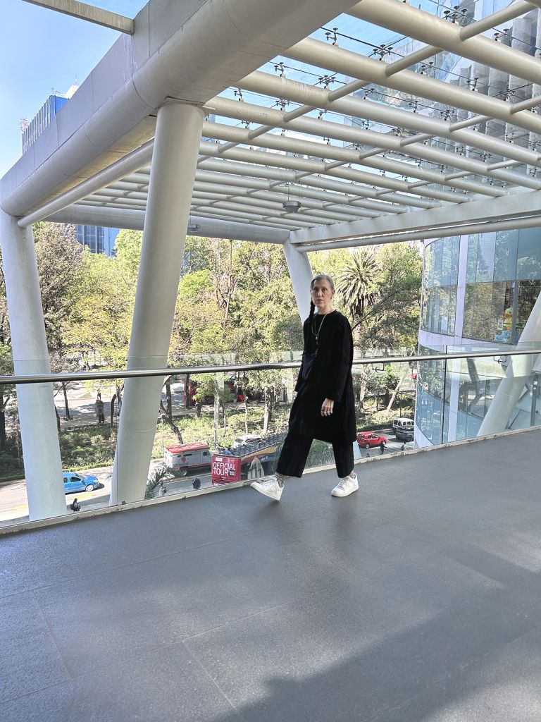 Person in black outfit standing on modern architectural bridge or walkway with geometric white structure and urban skyline background.