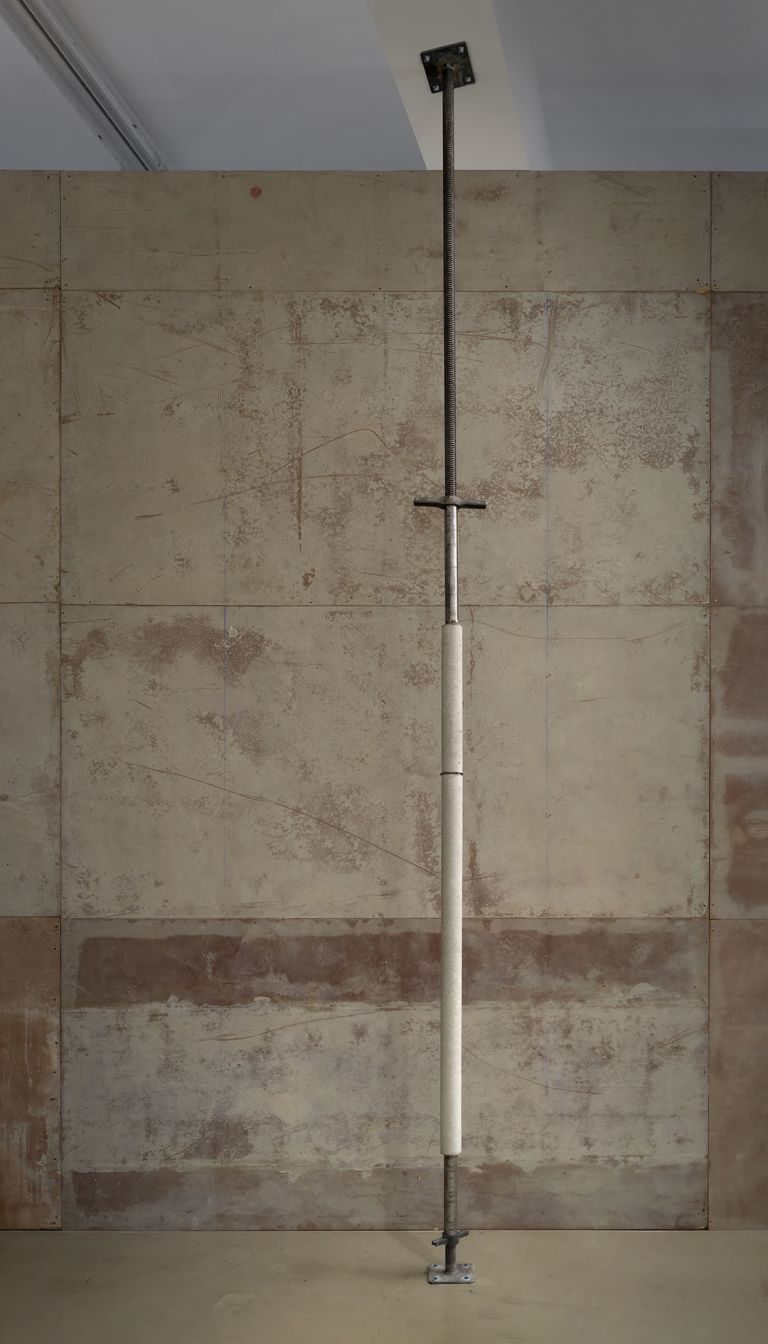 Floor-to-ceiling metal pole or rod installation against concrete wall panels in gallery space.