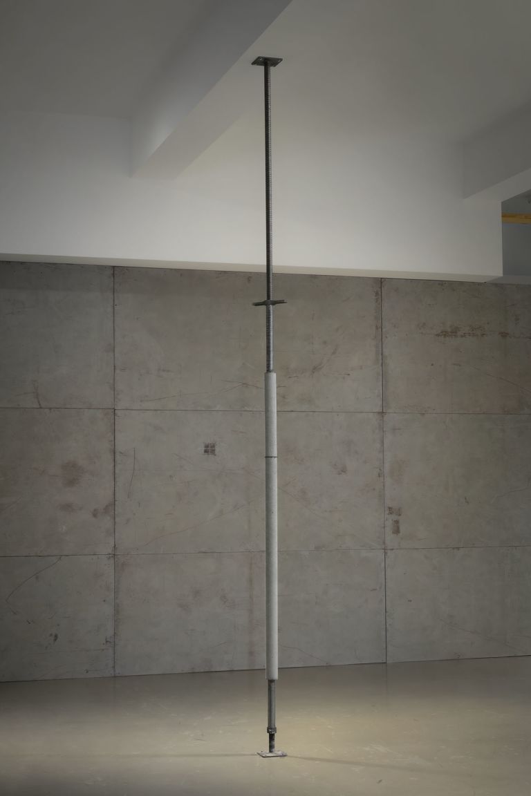 Vertical metal pole extending from floor to ceiling in minimalist gallery with concrete wall panels.