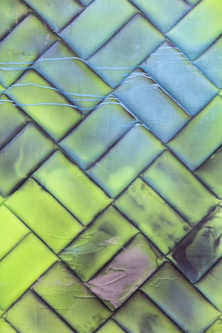 Painted mattress artwork with green and blue woven basketweave pattern and dark elements at bottom, mounted on white wall.