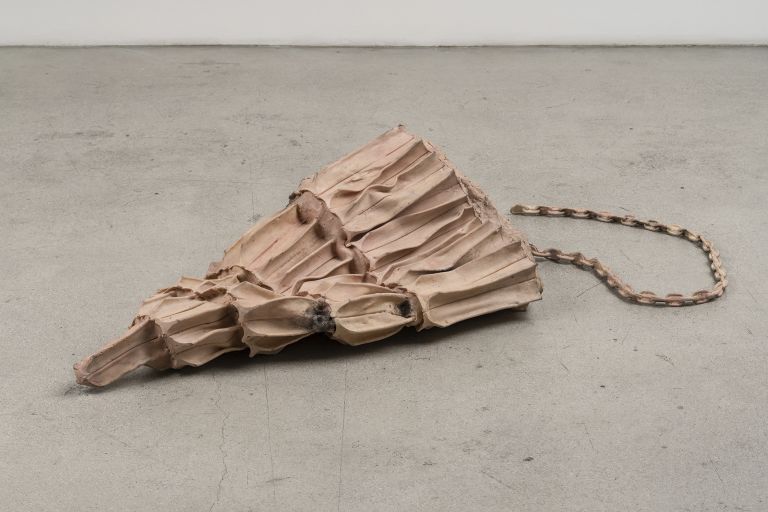 Sculptural object resembling layered fabric or paper with braided rope attachment on concrete floor.