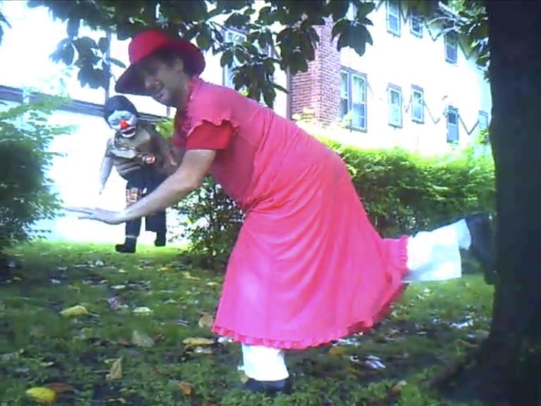 Person in bright pink dress and hat playing with child in grassy yard beside brick building.