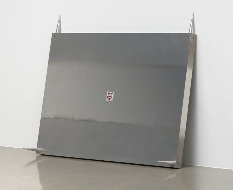 Minimalist installation featuring a large gray metal panel with two pointed spikes mounted on top and a small image in the center.