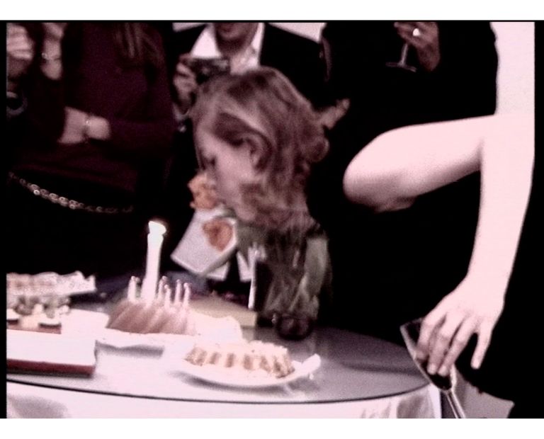 Vintage color film still showing a child blowing out candles on a birthday cake surrounded by hands and figures.