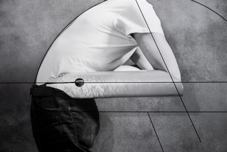 Person in white shirt and dark pants standing against gray backdrop with geometric line overlay resembling golden ratio