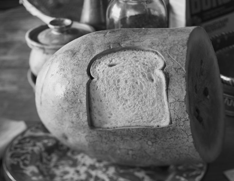 Black and white photo of a watermelon with slice of bread shaped cut-out on its surface, all surrounded by kitchen items including jars, cereal box, and plate.