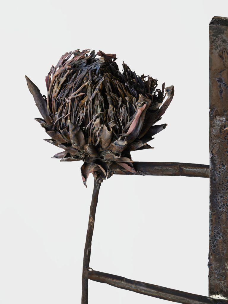Metal sculpture with two dried artichoke heads on crossbar, standing on conical base