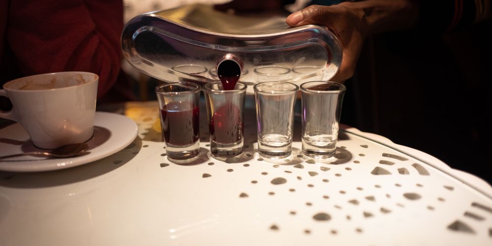 A flask, about 10 times the size of a regular flask pouring a deep red liquid into shot glasses.