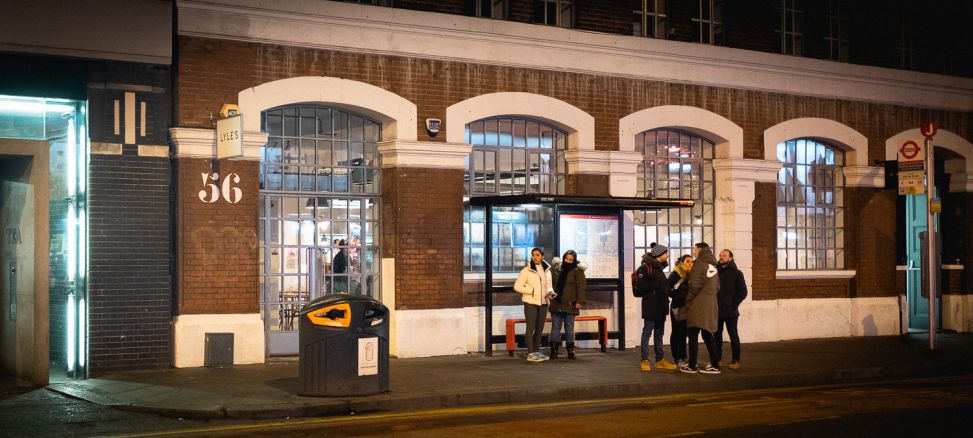 People standing at a bus stop that sits right in front of the brick building.