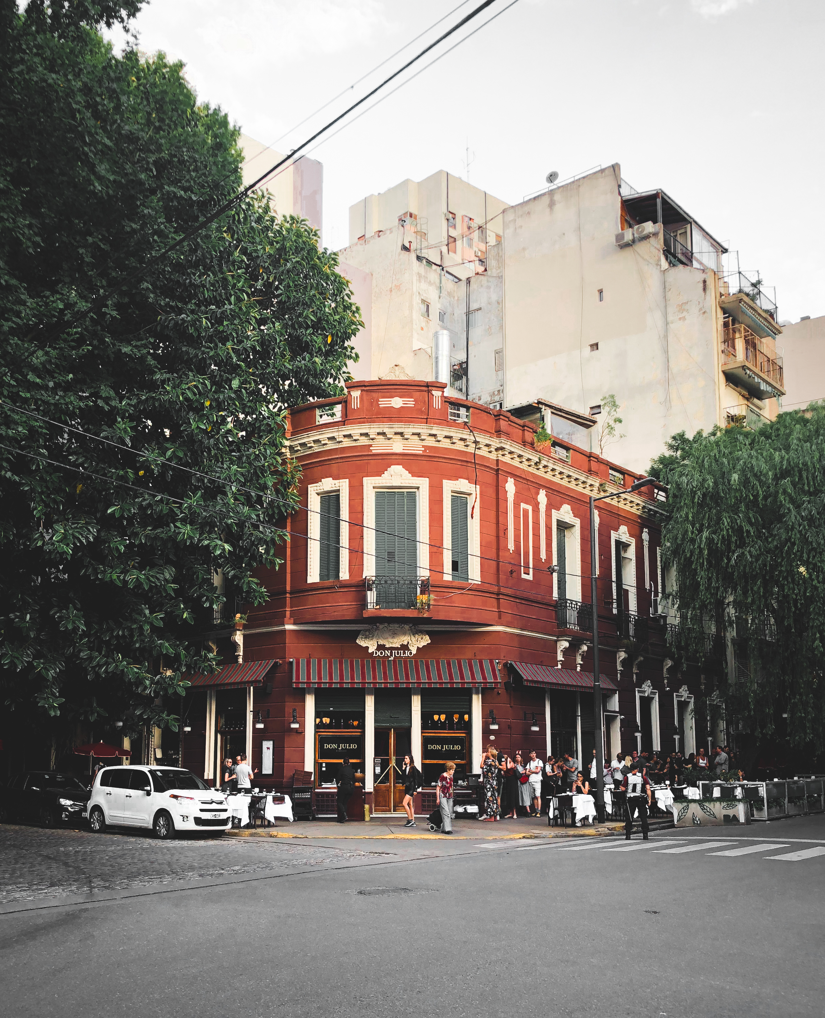The corner of a red brick building two stories high flanked by tree-lined streets