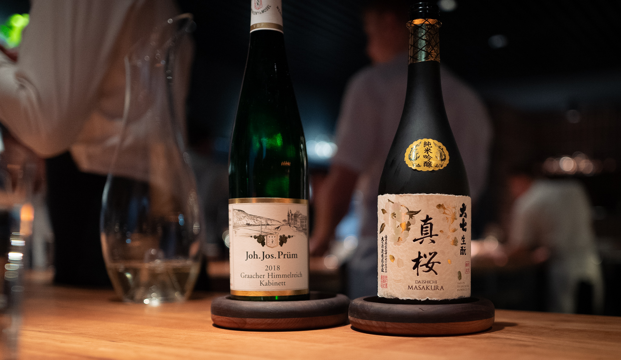 Two bottles of alcohol sit on coasters with chefs working away in the background.