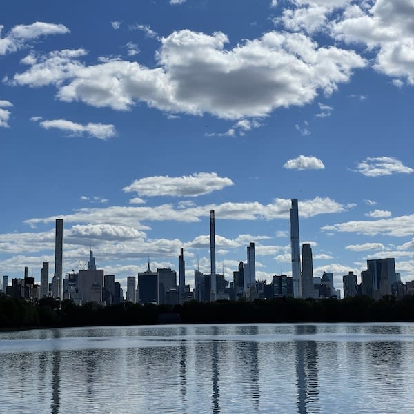 City skyline with tall skyscrapers reflected in a lake beneath a bright blue sky with scattered clouds