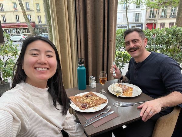 Two people smile for a selfie while dining at a Parisian café, with crêpes, drinks, and a city street visible outside.