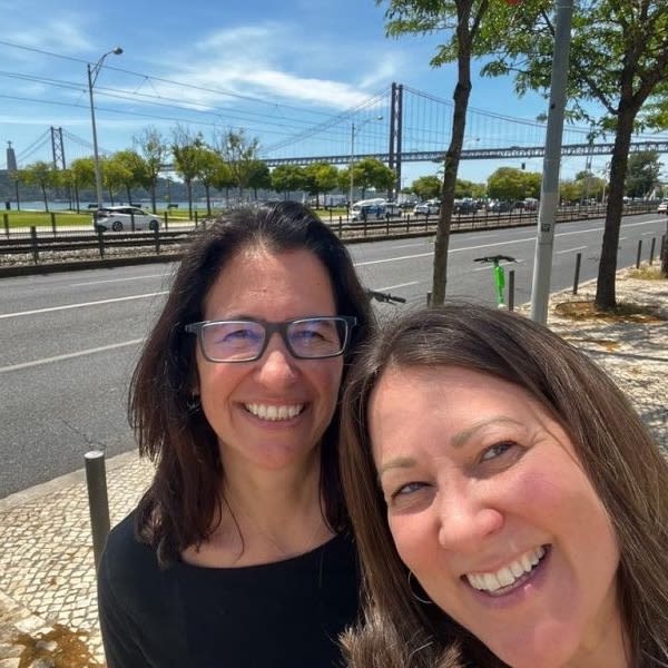 Two smiling women take a selfie on a sunny Lisbon street with the 25 de Abril Bridge visible behind them.