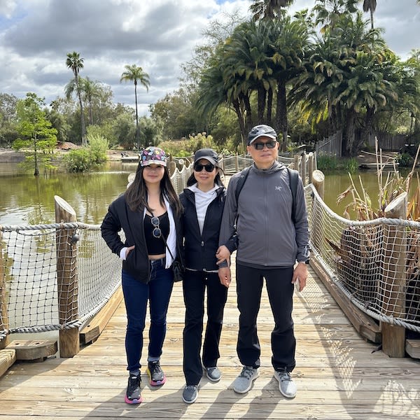 Three people pose on a wooden footbridge over a pond, surrounded by palm trees and lush greenery.