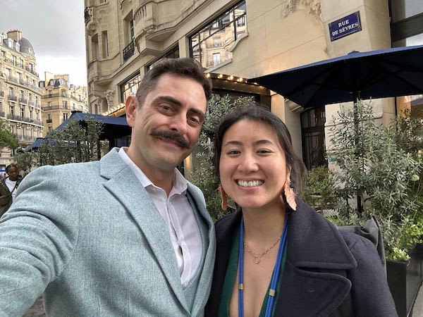 Two smiling people pose outside a Paris café beneath a Rue de Sèvres street sign.