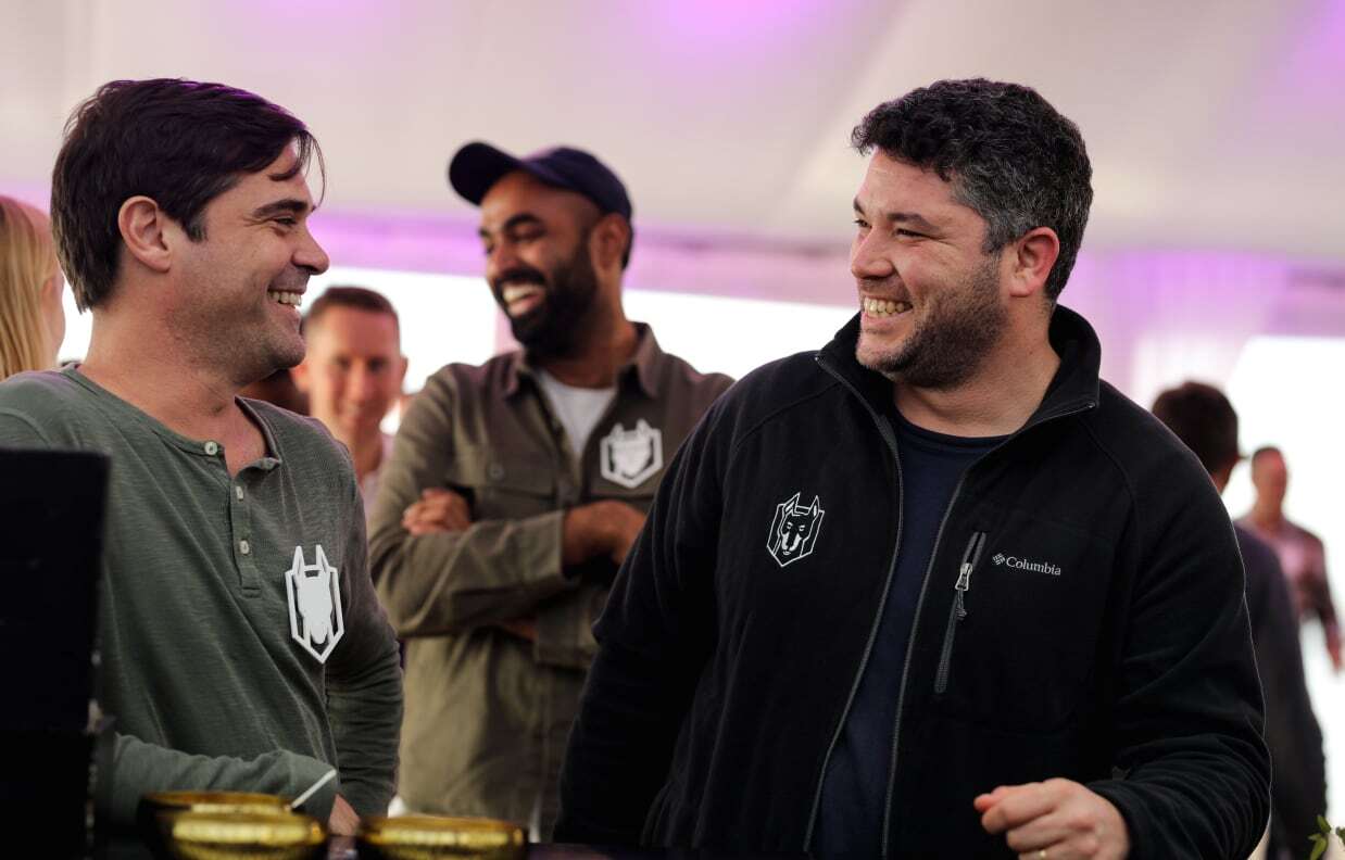 Dos hombres sonrientes conversan en un evento social en interiores mientras otro hombre está de pie detrás de ellos.