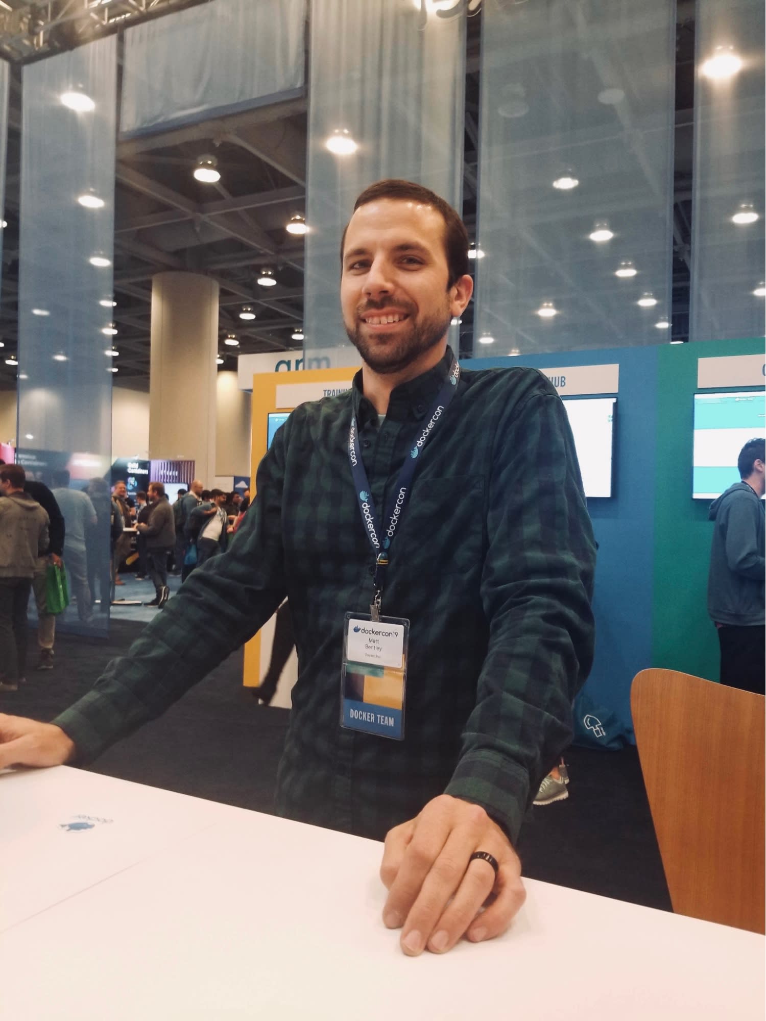 Matt Betley em pé atrás do balcão de um estande na DockerCon 2019, com as mãos apoiadas no balcão e usando um crachá com os dizeres "Docker Team"