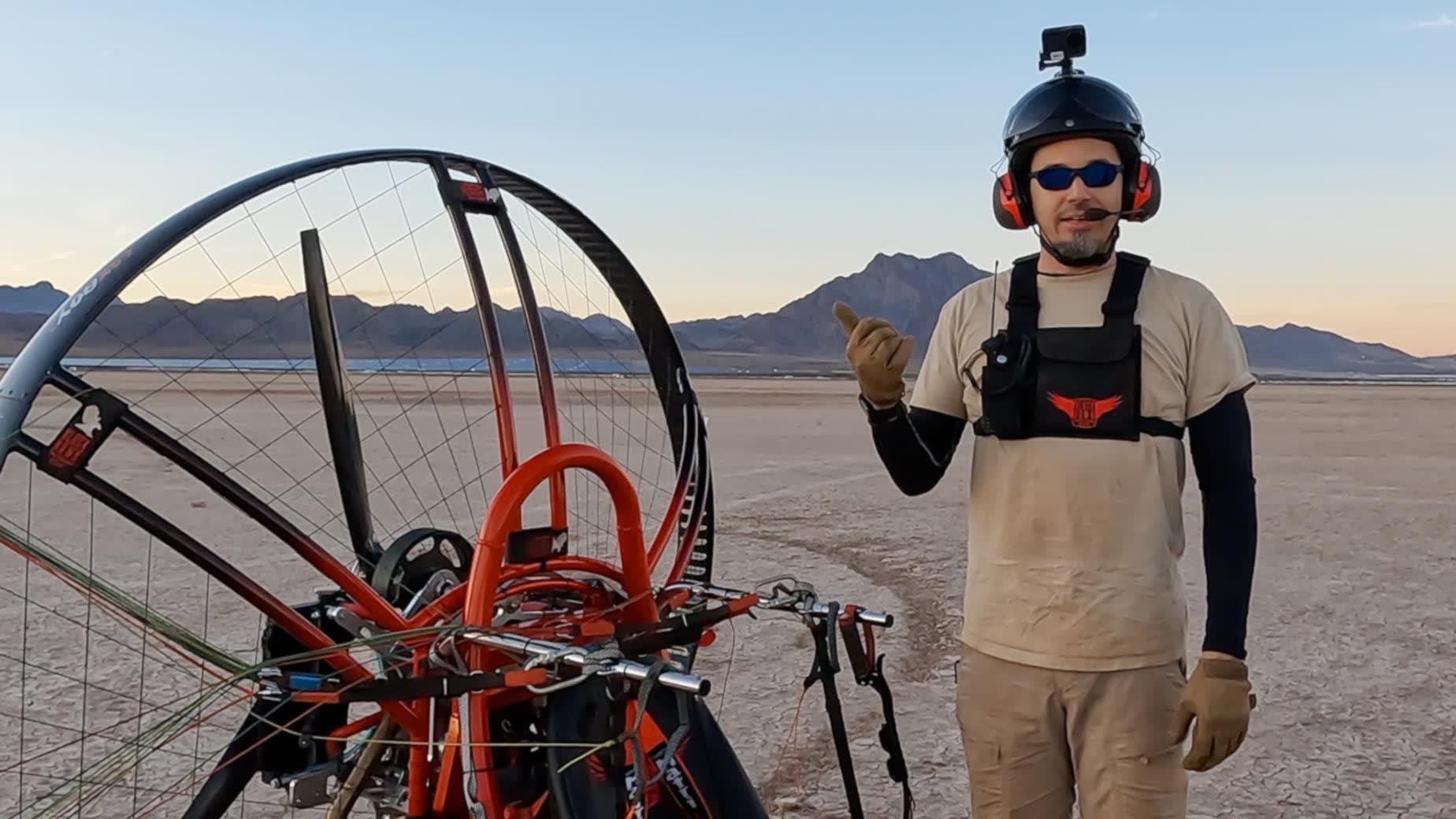 Adrian Goins usando capacete e com vários equipamentos de câmera, ao lado de uma aeronave paramotora quadricóptero laranja e preta
