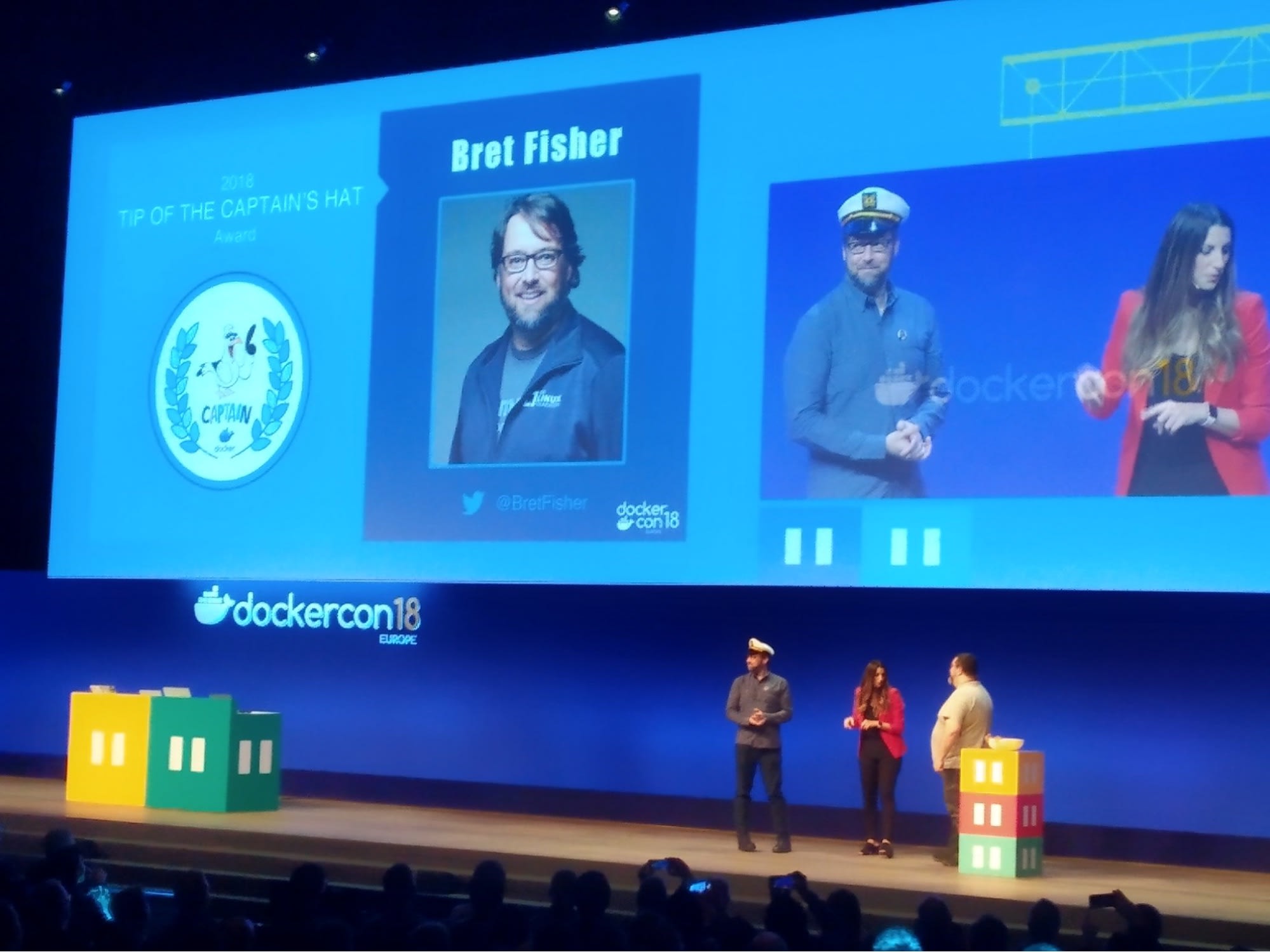 Palco da palestra principal da DockerCon 18 Europe, com Jenny Burcio e Mano Marks entregando a Bret Fisher o prêmio “Top of the Captains Hat”. Bret está usando um quepe branco de capitão de barco.