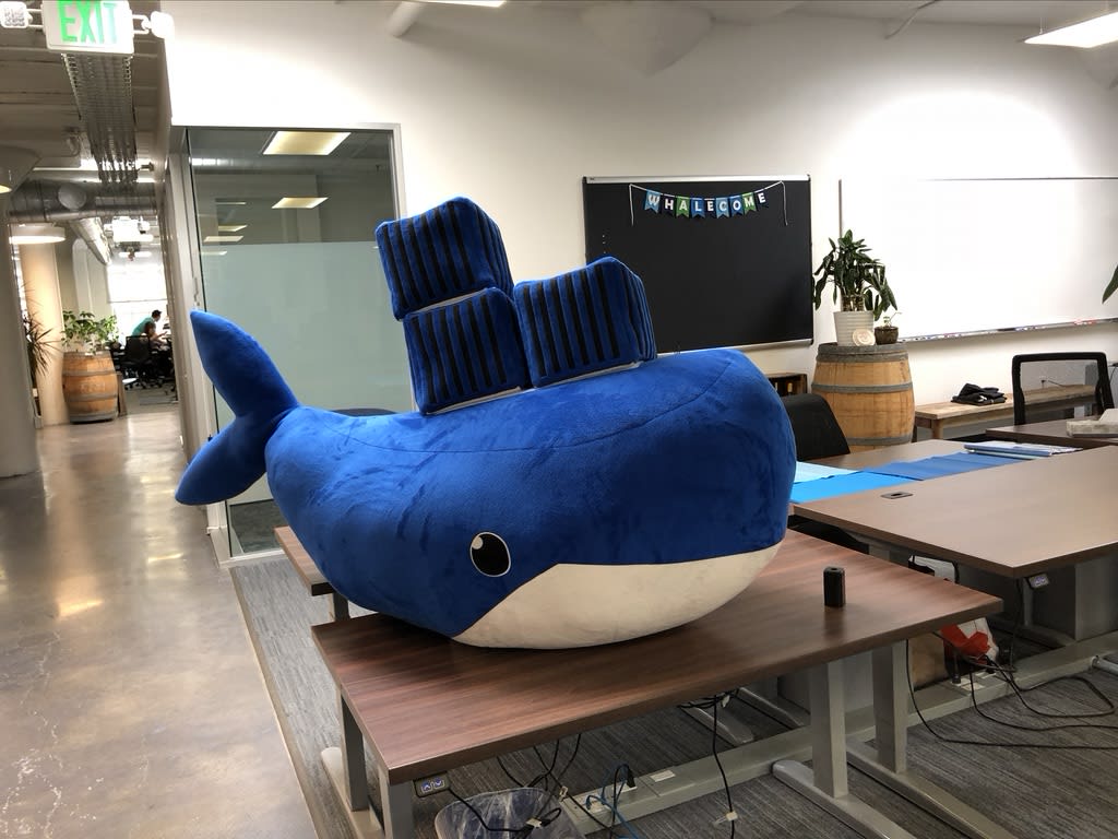 Um enorme (com mais de 1,5 m de comprimento) boneco de pelúcia azul do Moby, a Baleia, sentado em uma mesa de escritório vazia. Ao fundo, há uma faixa com a palavra “Whalecome” acima de um quadro de avisos vazio.