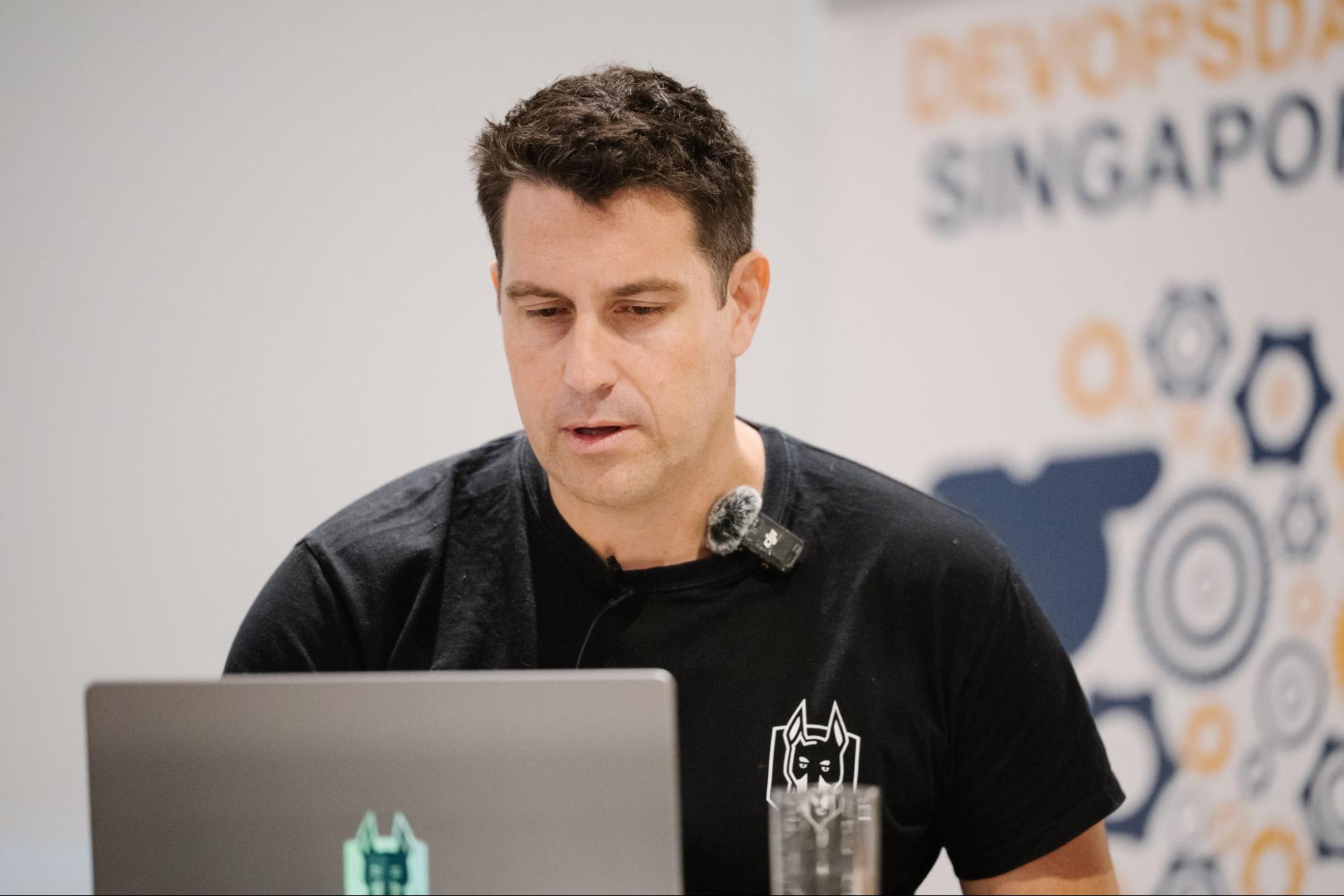 Speaker in a black shirt presenting behind a laptop, with a microphone and DevOpsDays Singapore backdrop