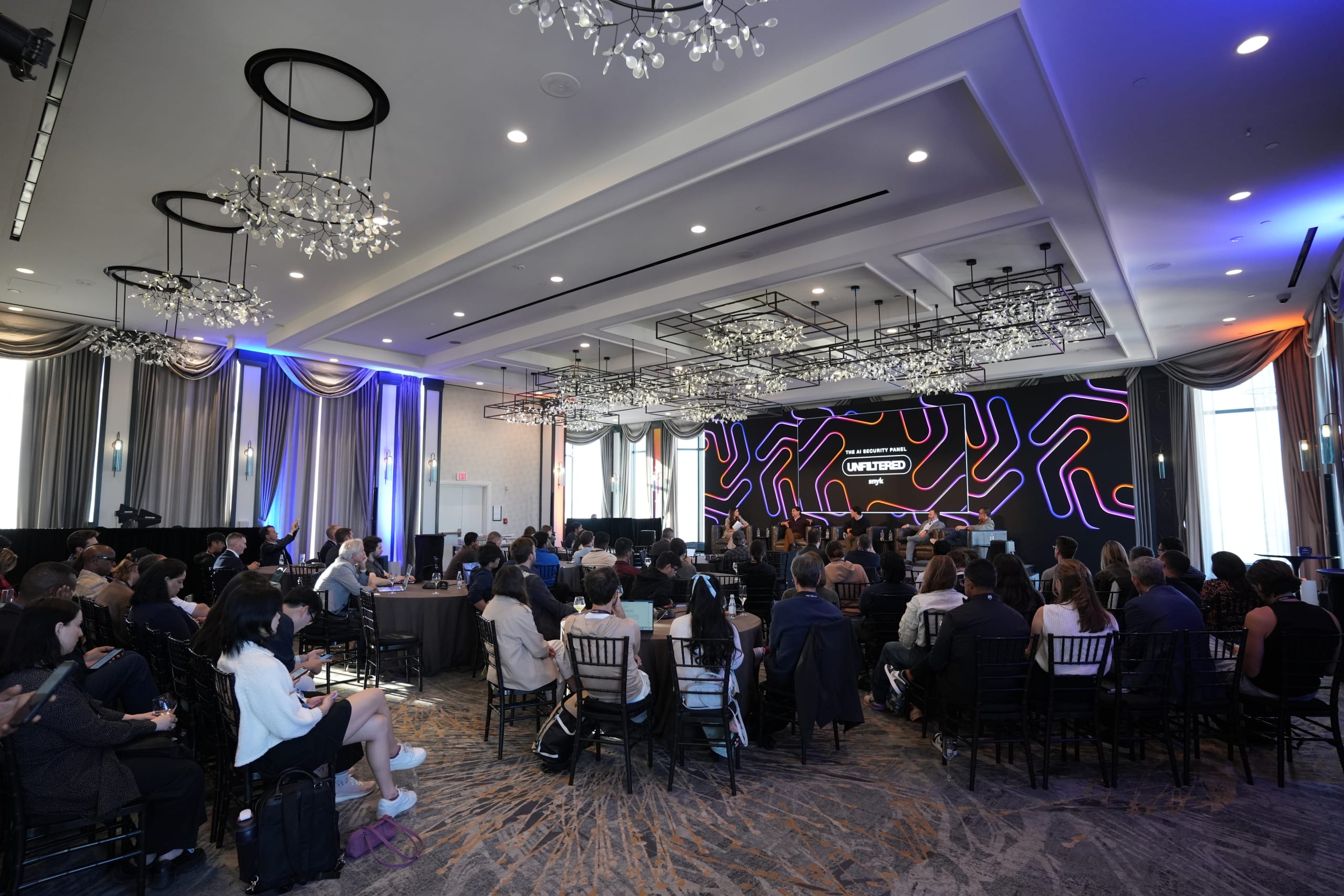 Audience seated at a conference panel beneath chandeliers, with a stage screen reading “The AI Security Panel” and “Unleashed.”