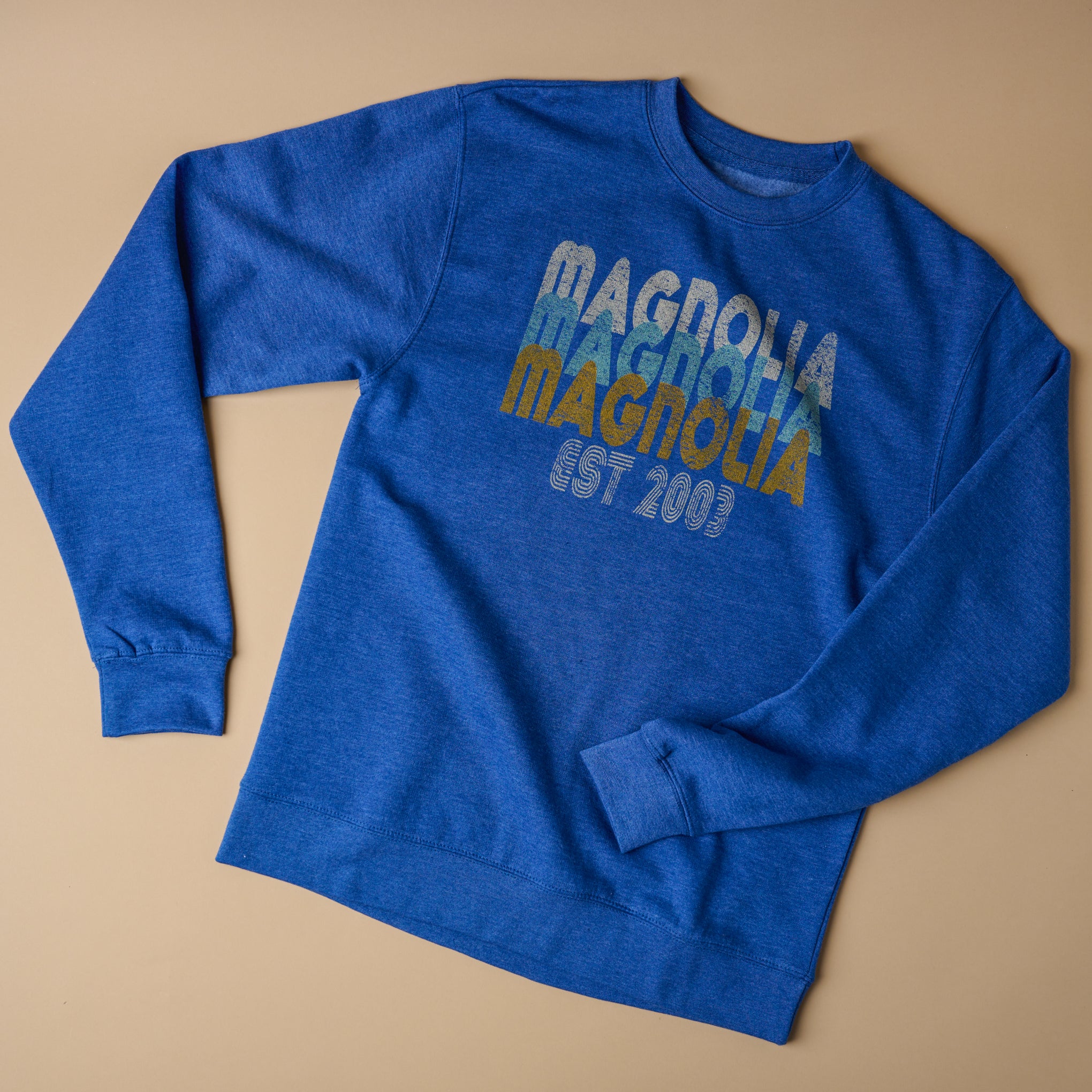 Royal Blue Magnolia sweatshirt reads "Magnolia" three times stacked on top of each other in different colors - white, light blue and yellow. Below that reads "Est. 2003" in white curved font.  On sale for $32.00, discounted from $64.00