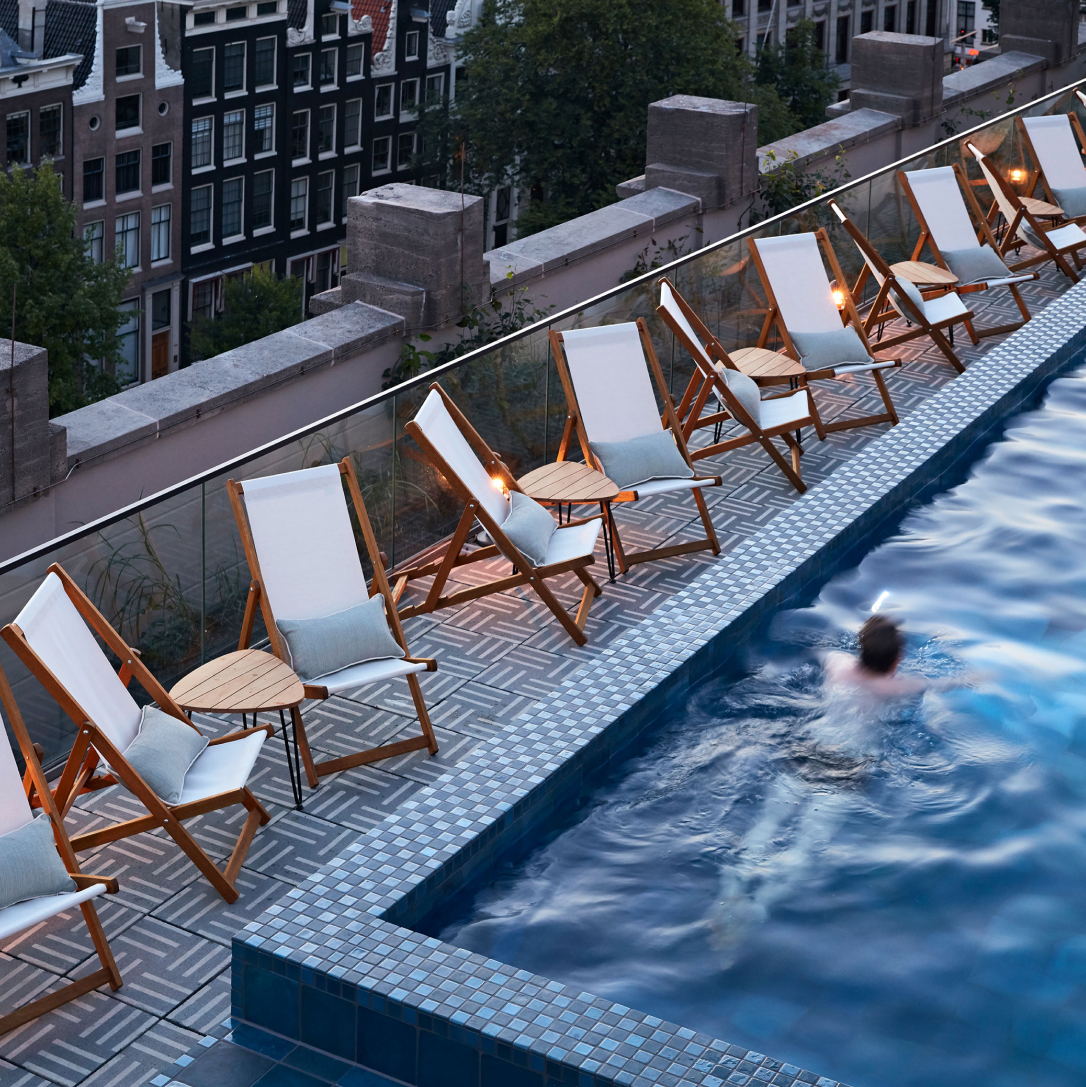 A person swimming in a rooftop pool surrounded by deckchairs with a city street below.
