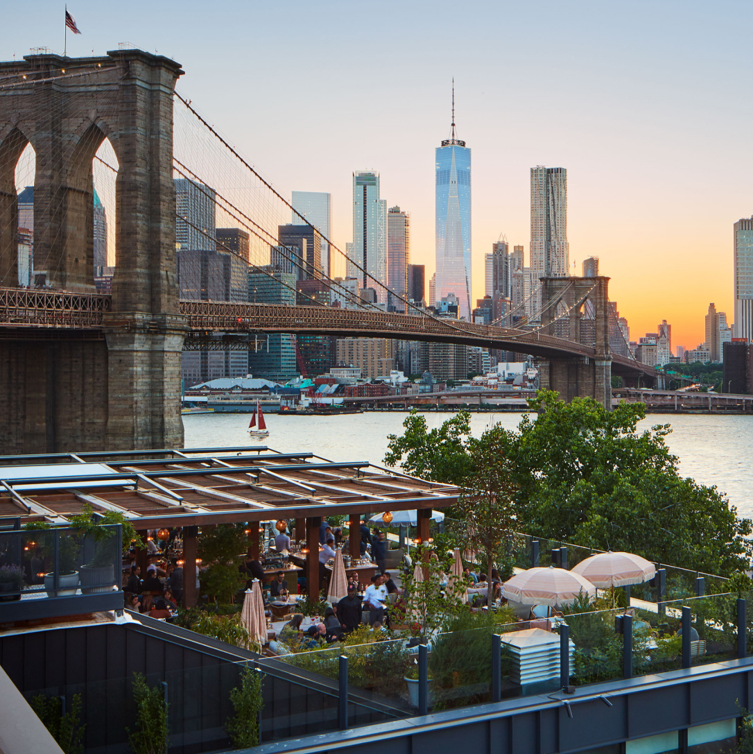A terrace overlooking a large bridge and river with a cityscape full of tall buildings in the background at sunset.