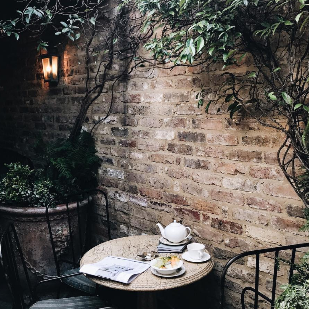 A outdoor terrace area with brick walls, plants and a table and chairs with a tea set laid on it.